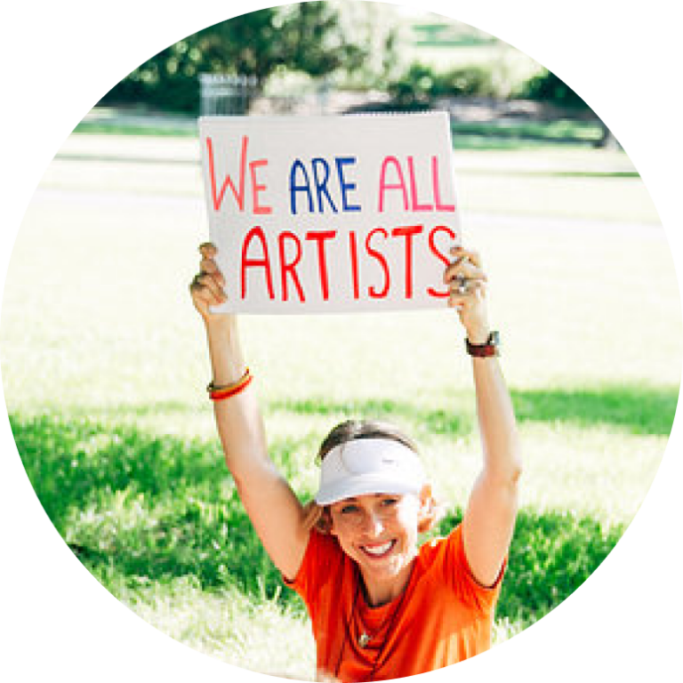 A woman (Robyn Katz) holding a handwritten sign that reads "we are all artists"
