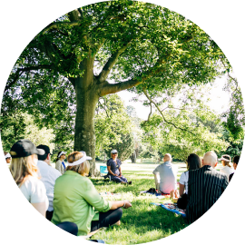 a group of women sitting outdoors in nature, under a large tree.